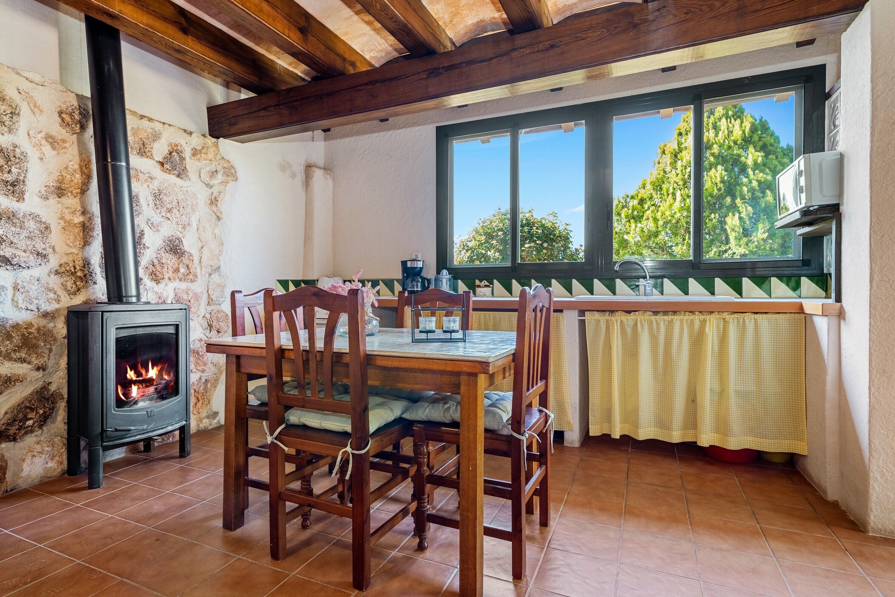 Une cuisine rustique avec un poêle à bois, une table à manger avec des chaises, une fenêtre donnant sur les arbres et un mur en pierre.