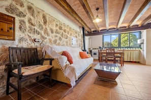 A cozy room with a stone wall, wooden furniture, a sofa with cushions, a glass coffee table, and a view of the outdoors through large windows.