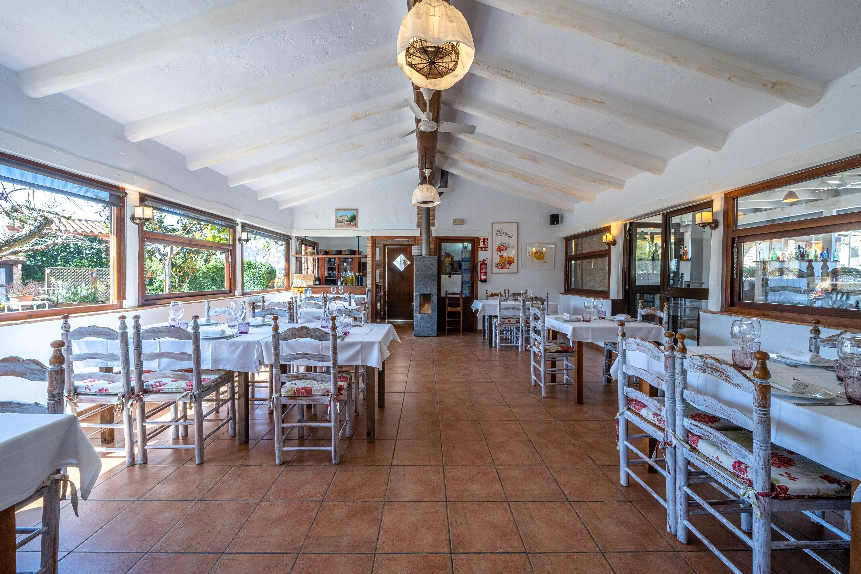 Un restaurante con mesas y sillas blancas, suelo de baldosas y un techo alto con vigas a la vista.