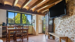 A rustic kitchen with wooden beams, a large window, a stone wall, and a flat-screen TV mounted on the wall. - Country House "Lo Paller" with Mountain View, Shared Pool & Wi-Fi (El Perelló)
