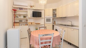 A compact kitchen with white cabinets, a round table with a pink tablecloth, a built-in TV, and a washing machine. - Apartment "Palau Corallo" with Shared Garden & A/C (Palau)