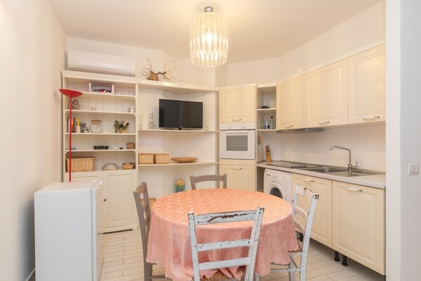 A compact kitchen with white cabinets, a round table with a pink tablecloth, a built-in TV, and a washing machine.