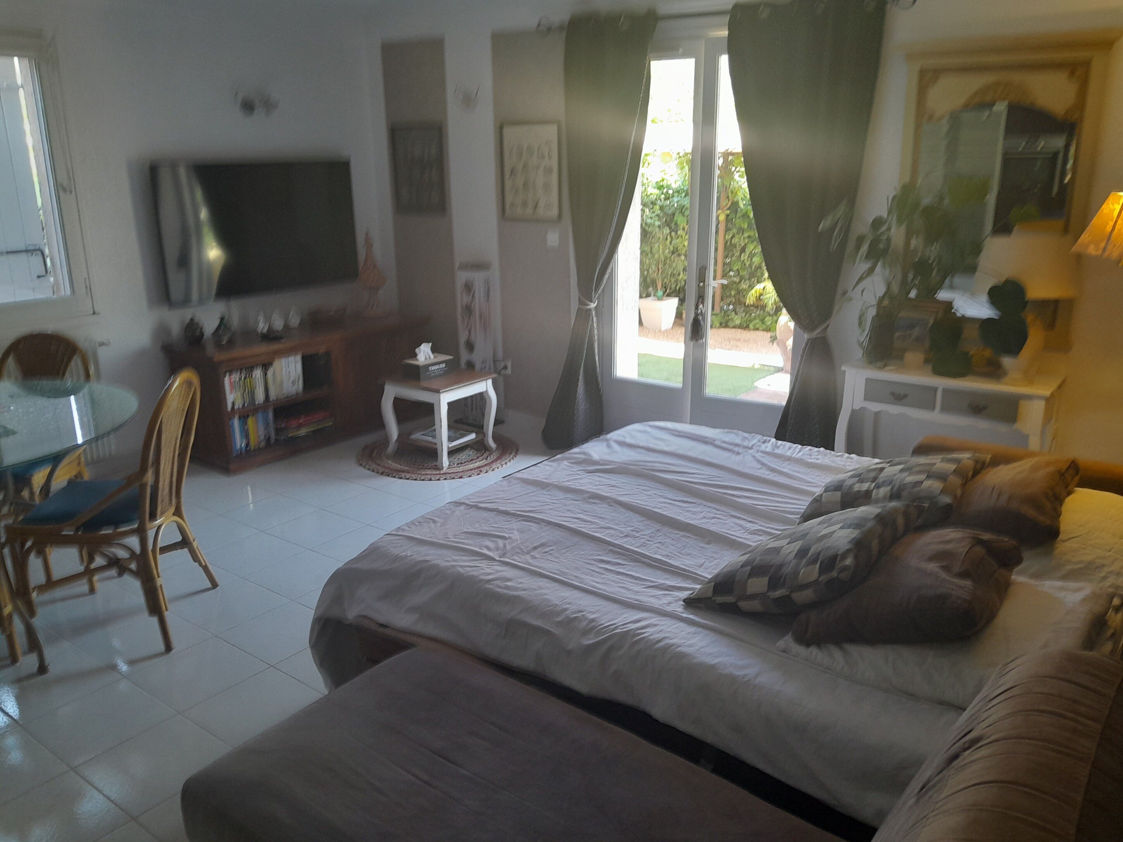 A bedroom with a bed, a TV, a dining table with chairs, and a small table with a lamp.