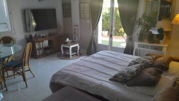 A bedroom with a bed, a TV, a dining table with chairs, and a small table with a lamp.