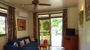 Ein Wohnzimmer mit blauem Sofa, Deckenventilator, Fernseher auf einem Holzkasten und einer großen Glasdoor zu einem Außenbereich.