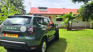 Ein grünes Auto vom Typ Dacia steht auf einer Wiese, im Hintergrund ist ein Gebäude mit rotem Dach zu sehen.