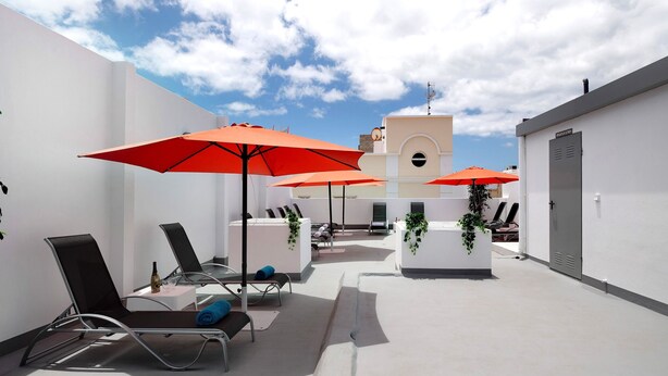Eine Dachterrasse mit Liegestühlen, roten Sonnenschirmen und einem weißen Gebäude mit einem Glockenturm.