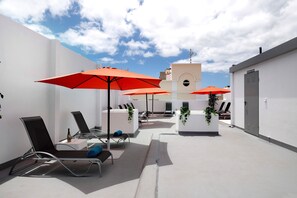 Una terraza en la azotea con sillones de descanso, sombrillas rojas y un edificio blanco con una torre del reloj.