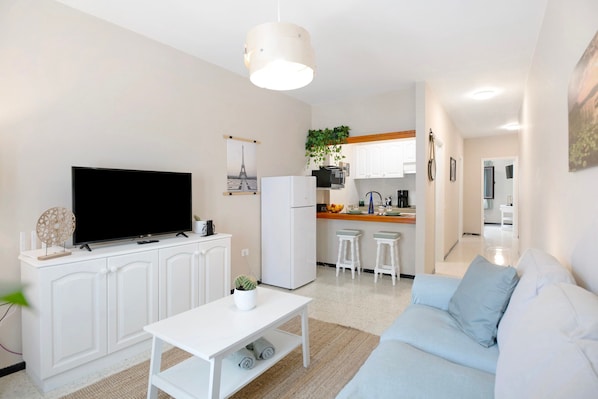 A modern living room with a flat-screen TV, white furniture, a kitchen area with bar stools, and a dining table.
