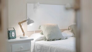 Ein ordentlich bezogenes Bett mit weißer Tagesdecke und Kissen, eine Nachttischlampe und ein Vintage-Radio auf einem weißen Nachttisch.