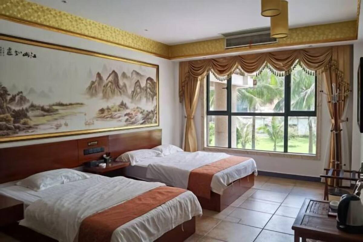 A hotel room with two beds, a large painting on the wall, a window with curtains, and a view of greenery outside.