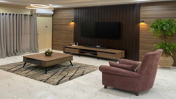 A modern living room with a flat-screen TV, wooden panel wall, and a plush armchair. - Integral Consults Apartment (Abuja)