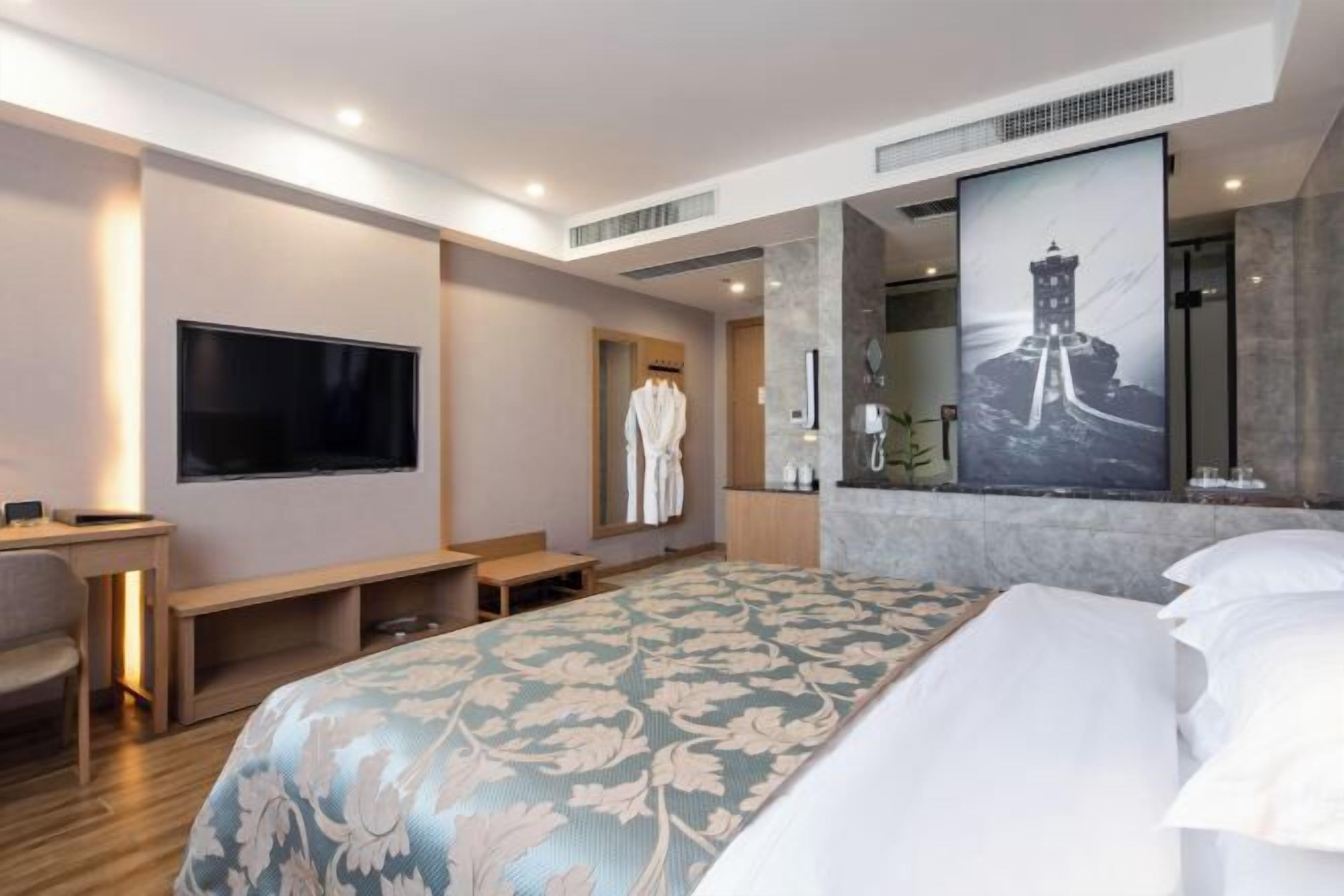A modern hotel room with a large bed, a flat-screen TV, a desk, and a bathroom with a large mirror and a framed picture of a lighthouse.