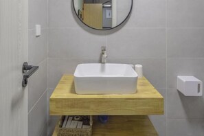 A bathroom with a rectangular white sink, a wooden countertop, a round mirror, and a towel dispenser. - Chaoqibanpo (Zhoushan)