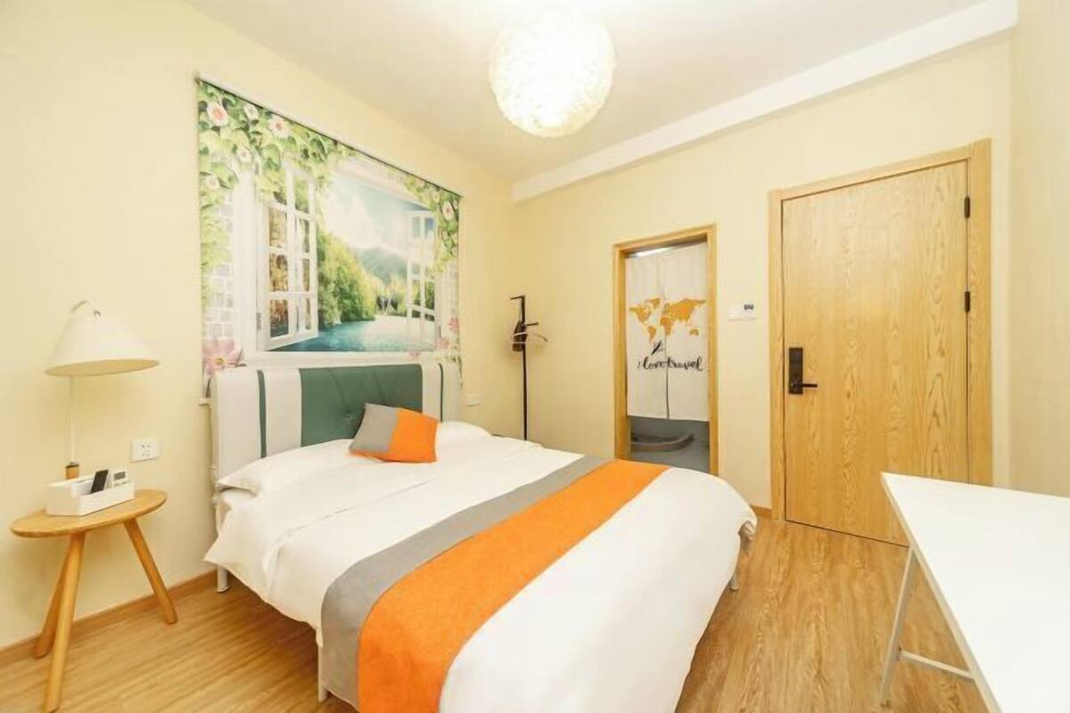 A hotel room with a bed, a small table, a lamp, a painting on the wall, and a wooden door.