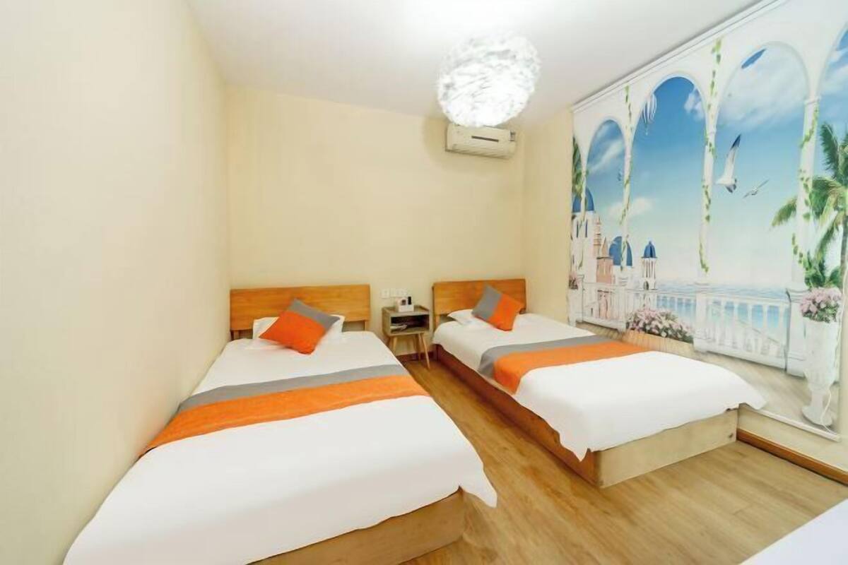 A hotel room with two beds, each with white bedding and orange accents, a wooden headboard, a nightstand, and a large window with a mural of a seaside scene.