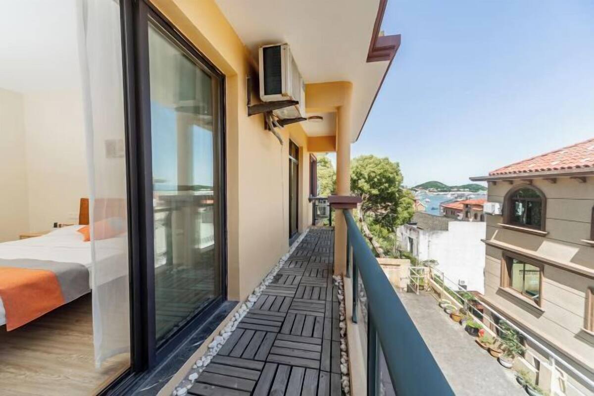 A balcony with a wooden floor, a bed with an orange blanket, and a view of the sea and buildings.