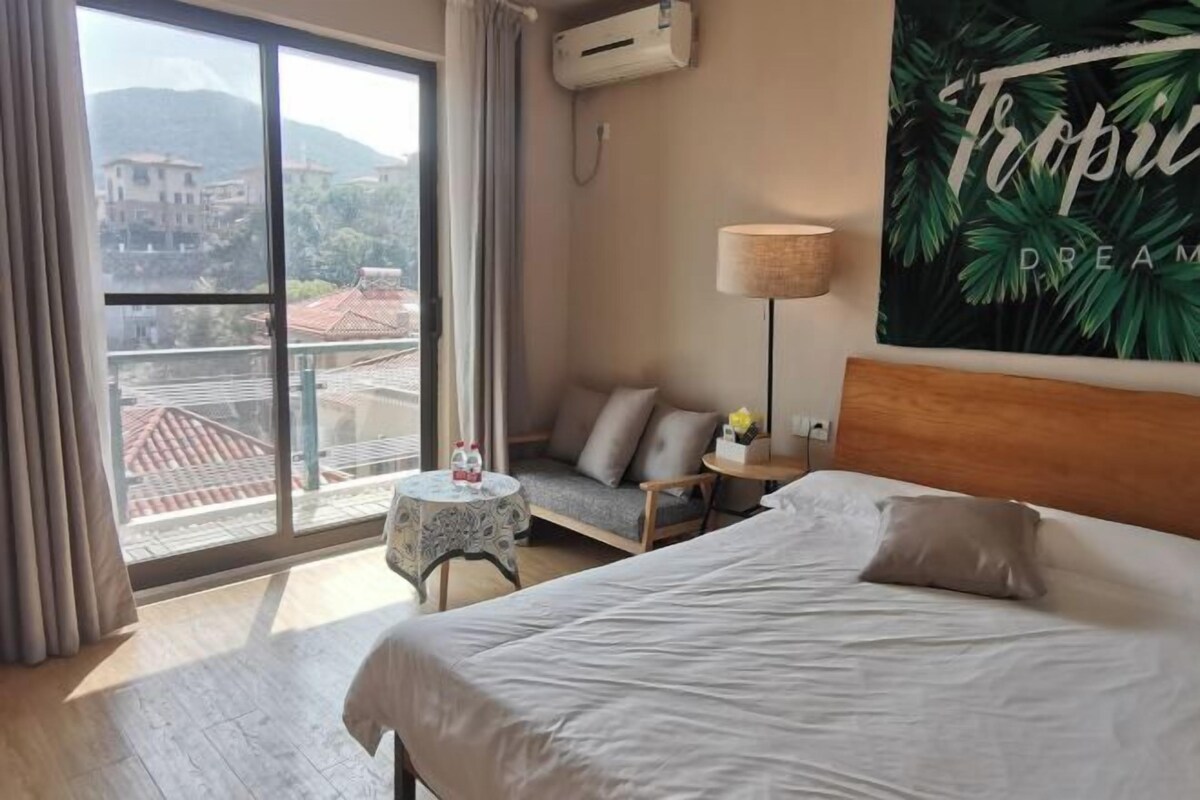 A hotel room with a large window, a bed, a bedside table, a lamp, and a tropical-themed wall art.