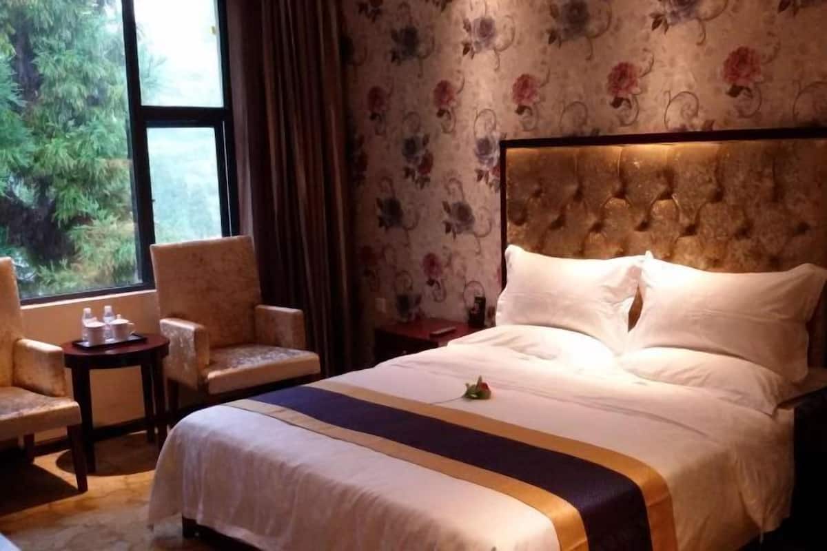 Quarto de hotel com uma cama grande, duas poltronas, uma mesinha com um tabuleiro e uma janela com vista para árvores.