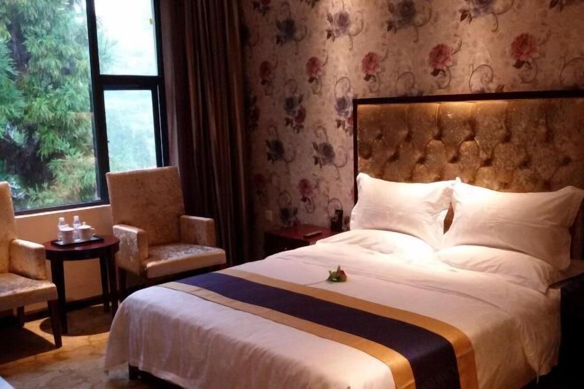 Habitación de hotel con una cama, dos sillones, una mesita y una ventana con vistas a árboles.