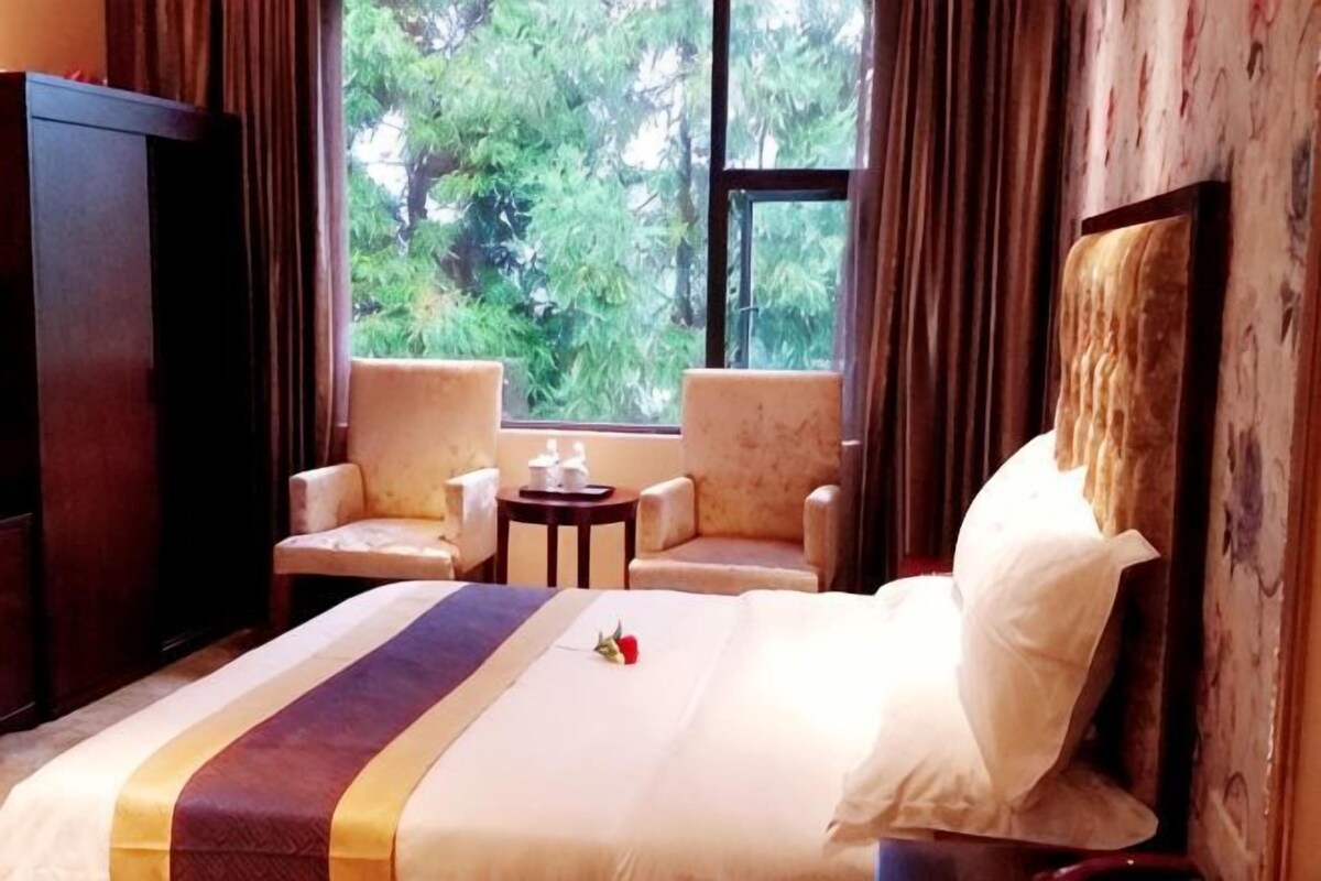 Habitación de hotel con cama, dos sillones, una mesita y ventana con vistas a árboles.