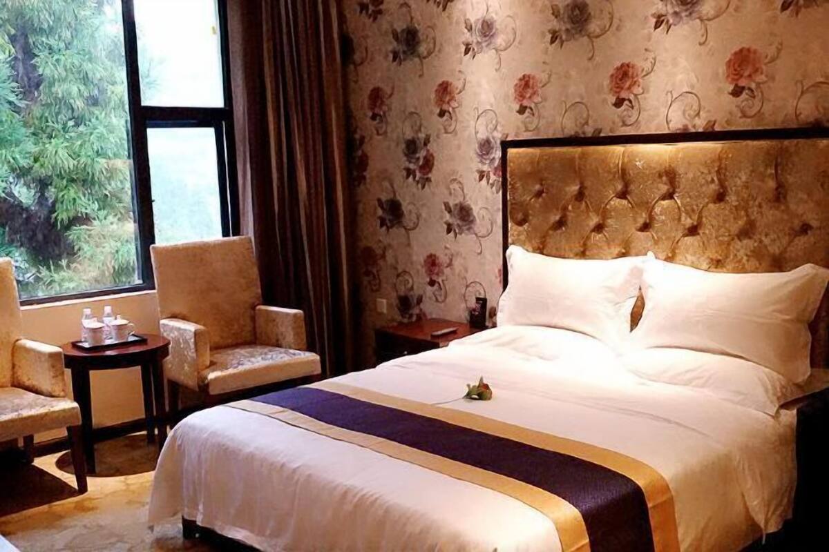 Quarto de hotel com uma cama, duas poltronas, uma pequena mesa com uma garrafa e uma janela com vista para árvores.