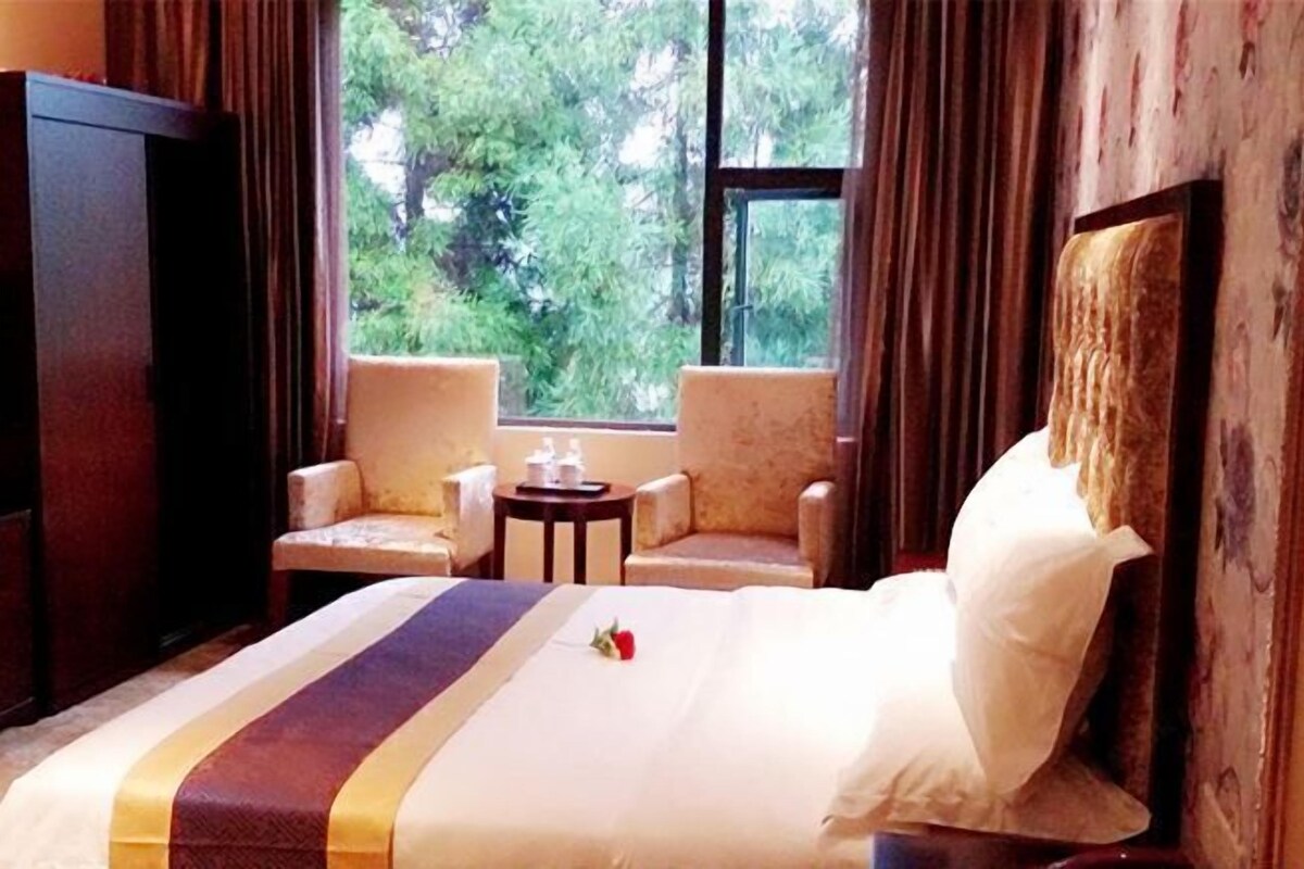 Habitación de hotel con una cama, dos sillones, una mesita y un ventanal con vistas a árboles.