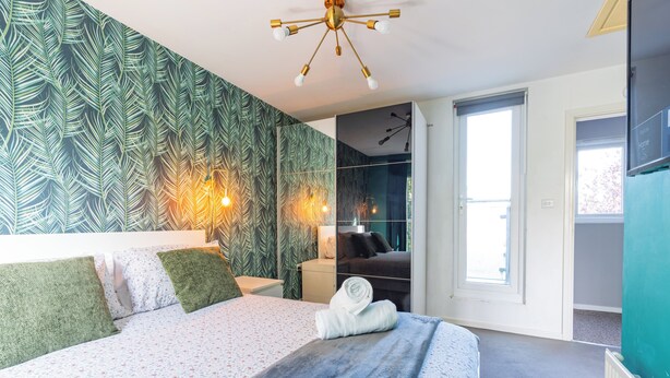 Une chambre moderne avec un grand lit, un mur orné d’un motif de feuilles vertes, un luminaire au plafond et une vue sur un balcon.