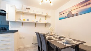 Un espace repas moderne avec une table en bois foncé, disposée pour quatre personnes, des meubles blancs et une étagère murale agrémentée d’objets décoratifs.