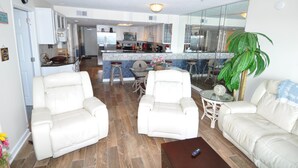A spacious living room with two white leather sofas, a glass coffee table, and a dining area with a small table and chairs.