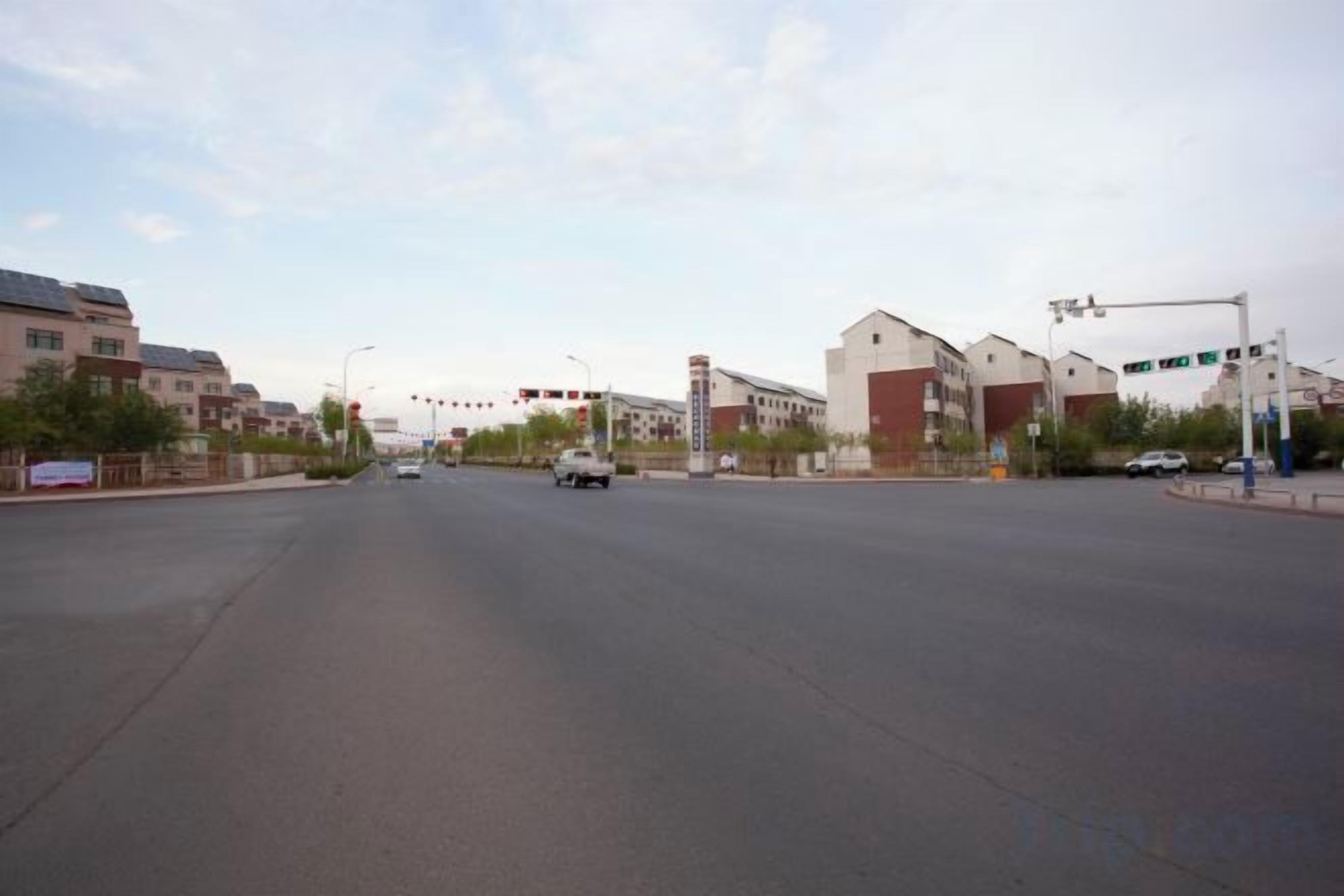 A wide urban road with buildings on both sides, traffic lights, and a clear sky.