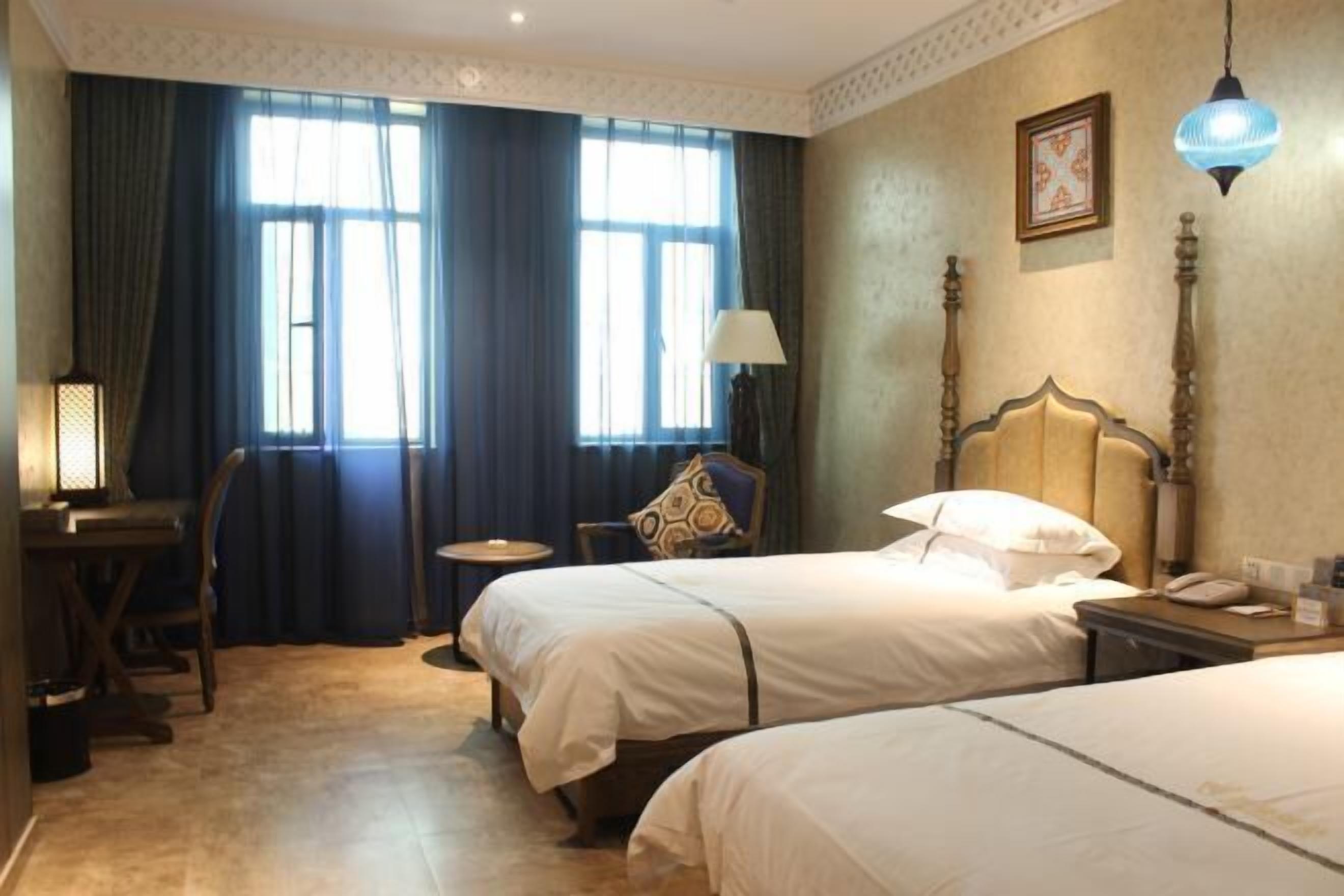 A hotel room with two beds, a desk, a chair, a lamp, and a bedside table.