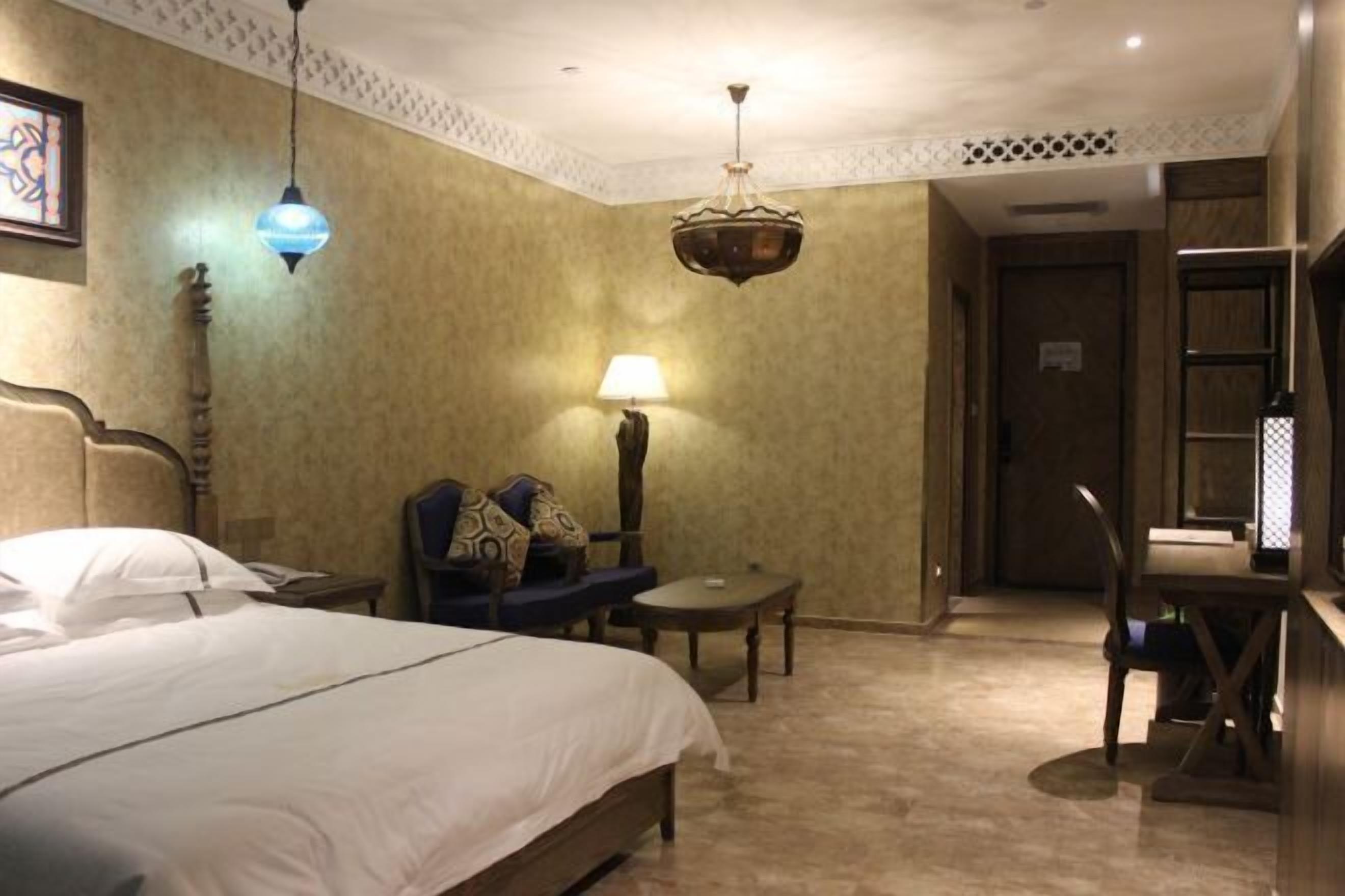 A hotel room with a bed, a desk, a chair, a lamp, and a hanging light fixture.