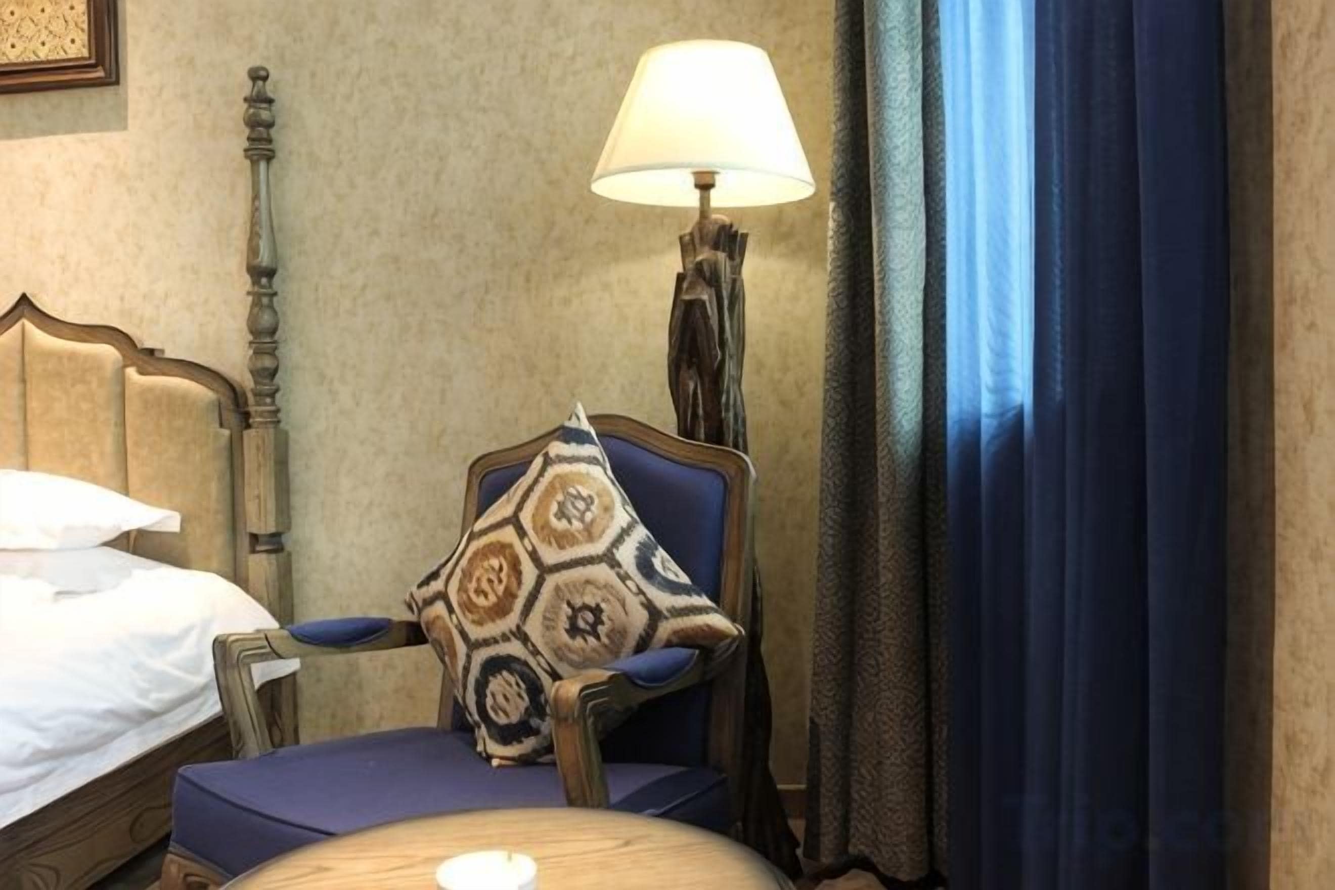 Habitación de hotel con cama, una silla con cojín estampado, lámpara y vistas al exterior.