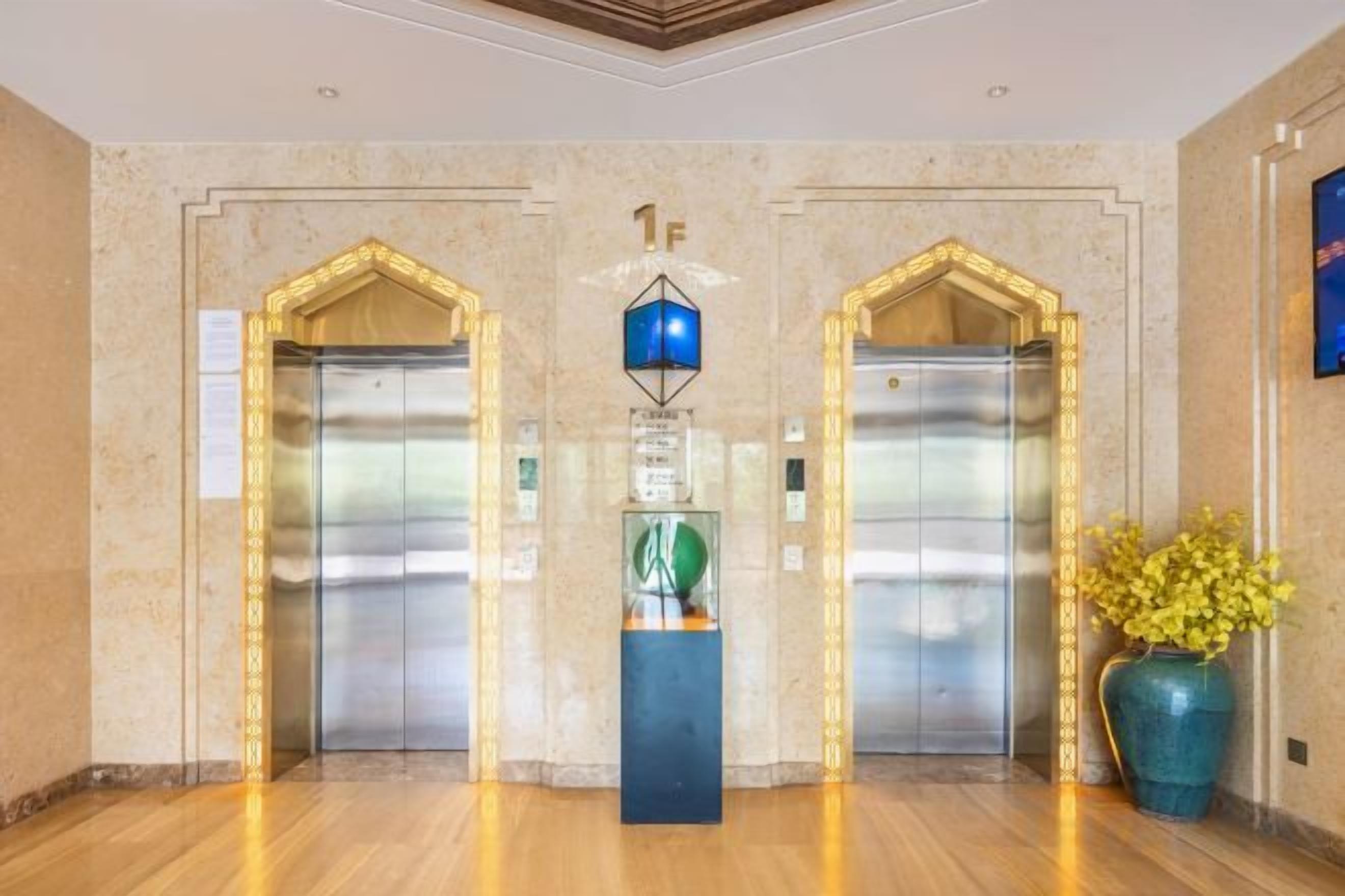 A lobby with two elevators, a blue decorative light fixture, and a vase with yellow flowers.