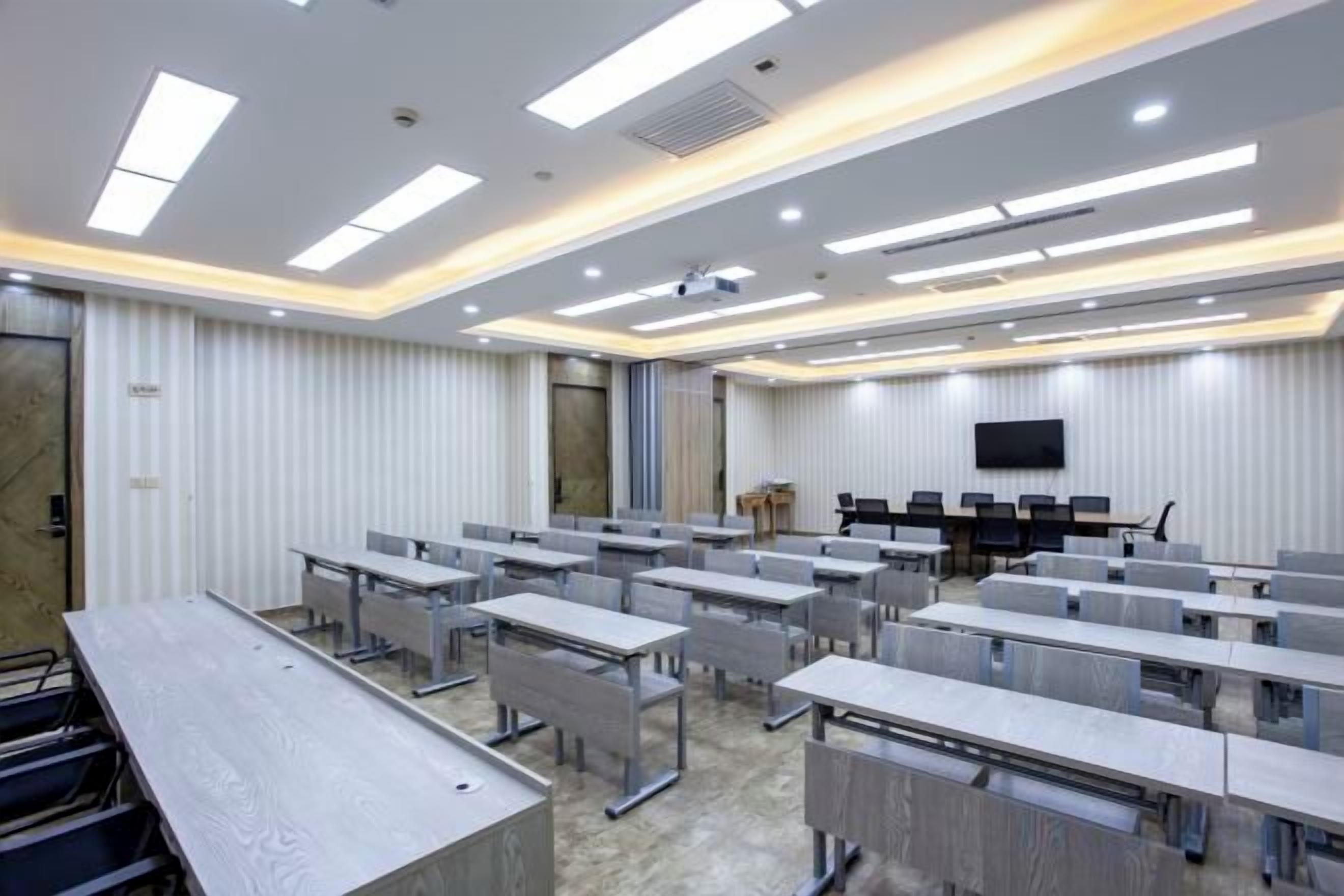 A conference room with rows of tables and chairs, a projector, and a flat-screen TV on the wall.