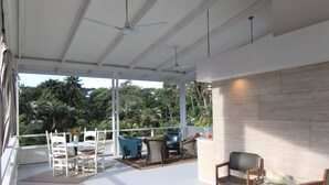 Un área de estar al aire libre con techo, con sillas y una mesa blancas, una mesa de vidrio con sillas, y vistas a vegetación exuberante.