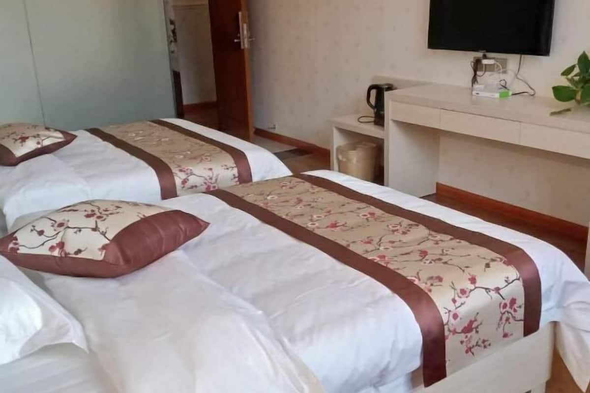 A hotel room with two beds, each with floral-patterned bedding and brown trim. There is a television mounted on the wall, a small shelf with a kettle and a plant, and a door leading to another room.