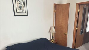 A single bed with a dark blue bedspread in a room with a wooden door, a small table with a lamp, and a framed picture on the wall.