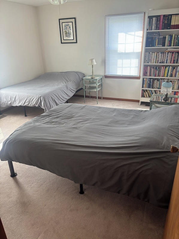 Un dormitorio con dos camas individuales, una mesita con lámpara, una estantería y una ventana con persianas.