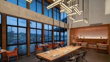 A modern dining area with a long table, multiple chairs, and a view through large windows.