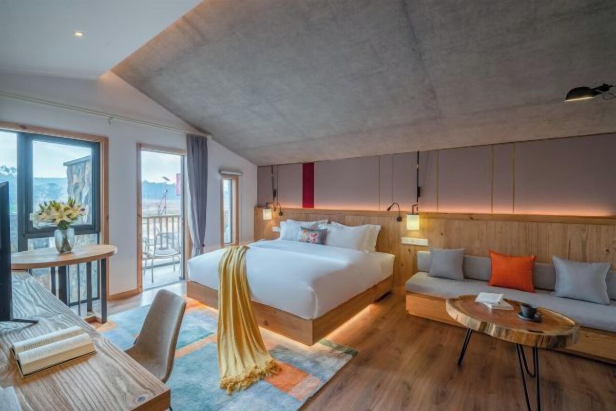 A modern hotel room with a large bed, a sofa, a small table, and a balcony with a view.