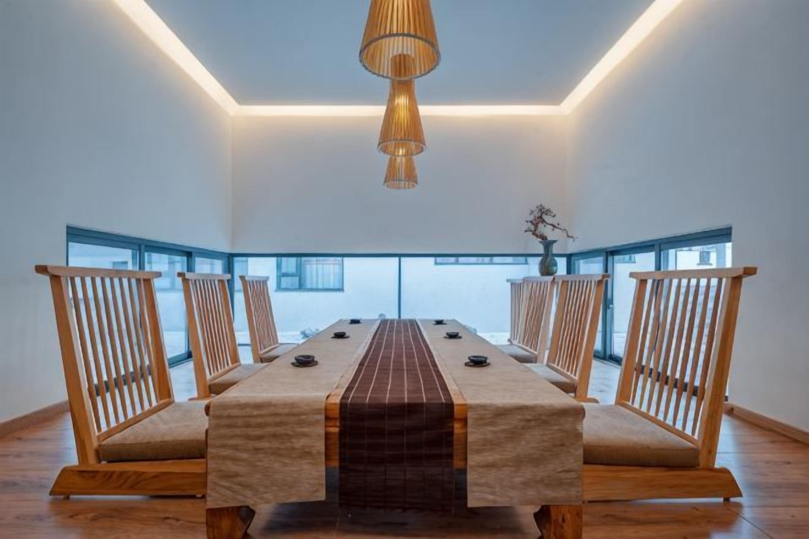 A long dining table with chairs, a hanging light fixture, and large windows.