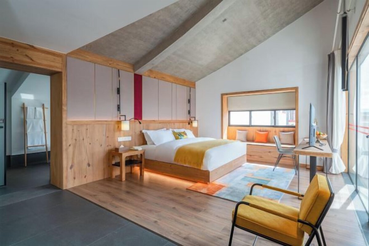 A modern bedroom with a sloped ceiling, a bed with a wooden headboard, a desk with a chair, and large windows.