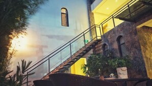 Una escalera con barandillas de vidrio que sube a un nivel superior, rodeada de plantas de interior y muebles de mimbre.