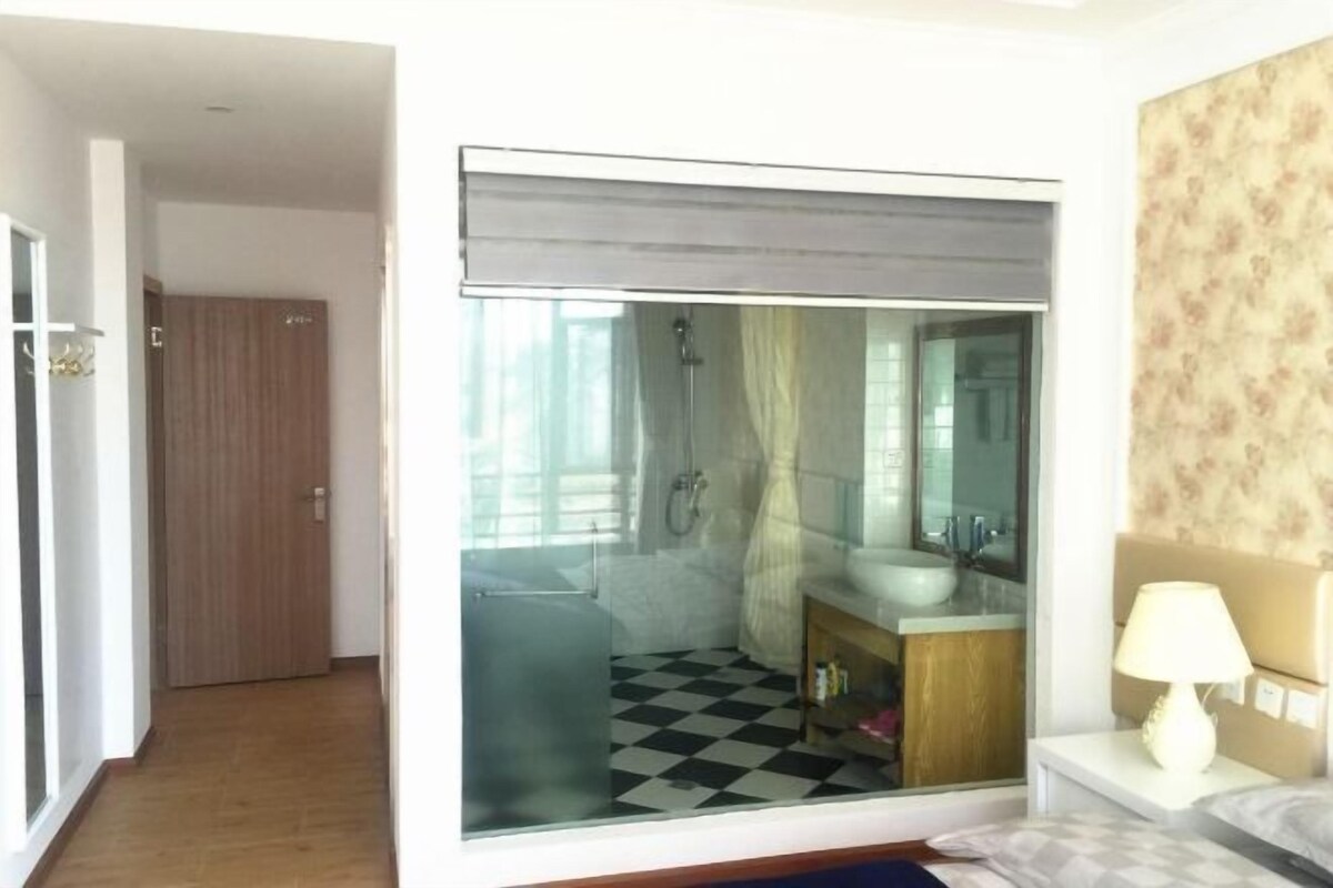 A hotel room with a checkered floor, a glass shower enclosure, a bedside table with a lamp, and a bed with a pillow.