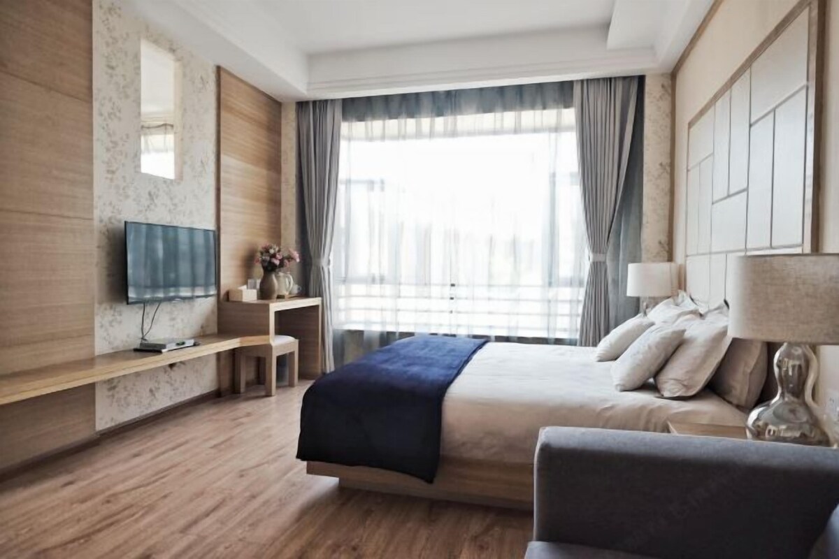 A modern hotel room with a bed, a TV, a desk, and a window with curtains.