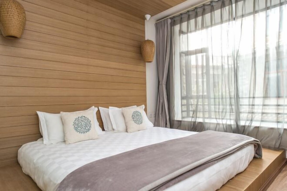 A bedroom with a wooden headboard, a large window with sheer curtains, and a bed with a white and brown blanket.