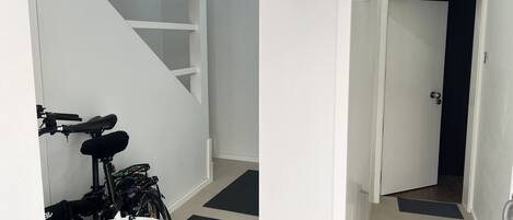 Un pasillo estrecho con una escalera blanca a la izquierda y una puerta al final. Una bicicleta está estacionada contra la pared.