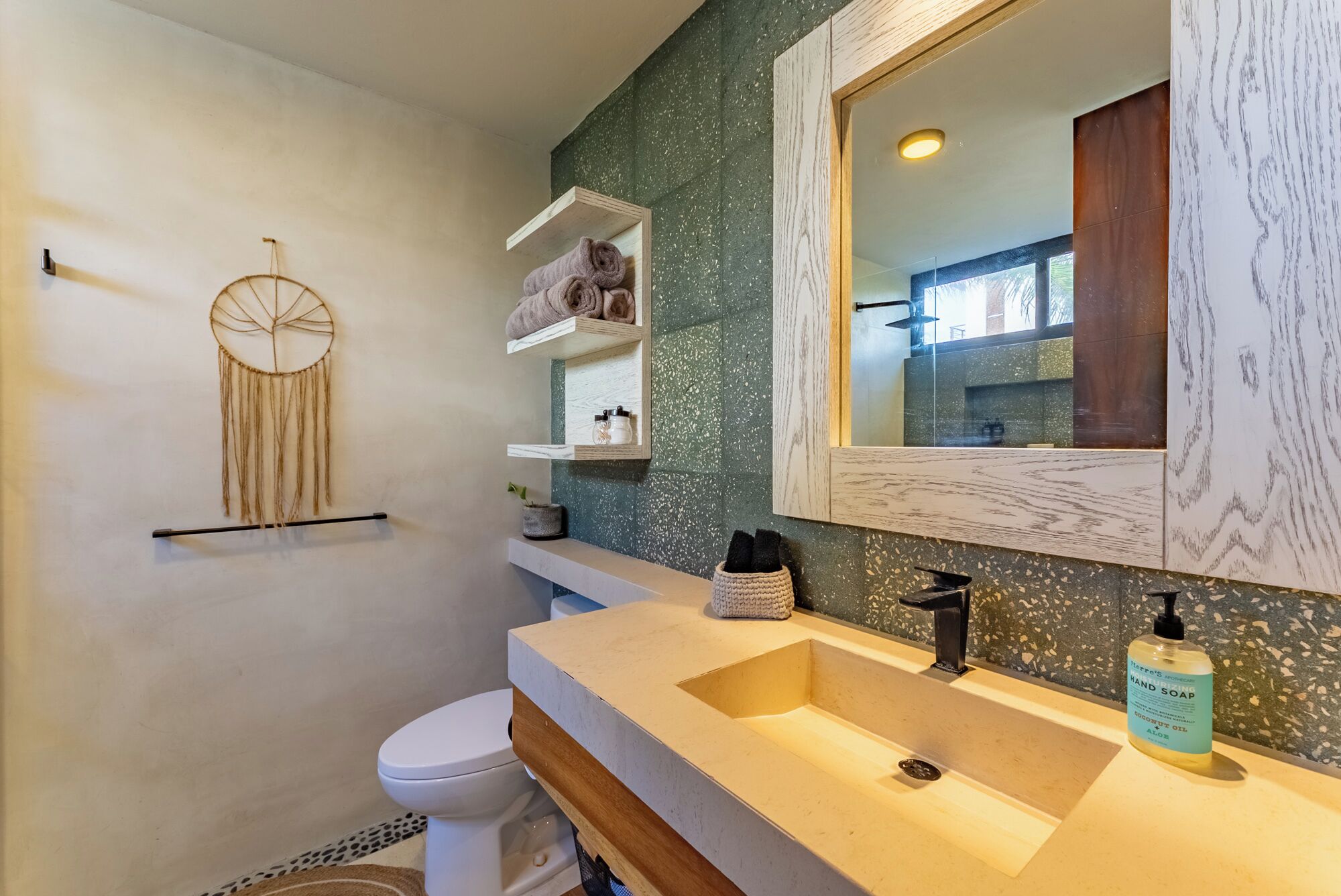 A bathroom with a white toilet, a beige sink with a wooden countertop, a mirror with a wooden frame, and shelves with towels and toiletries.